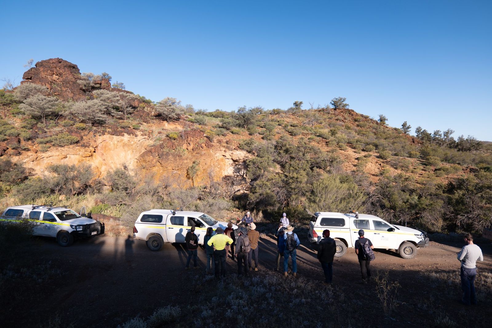The team gathered beside parked 4WDs below a rocky hill in late afternoon sun
